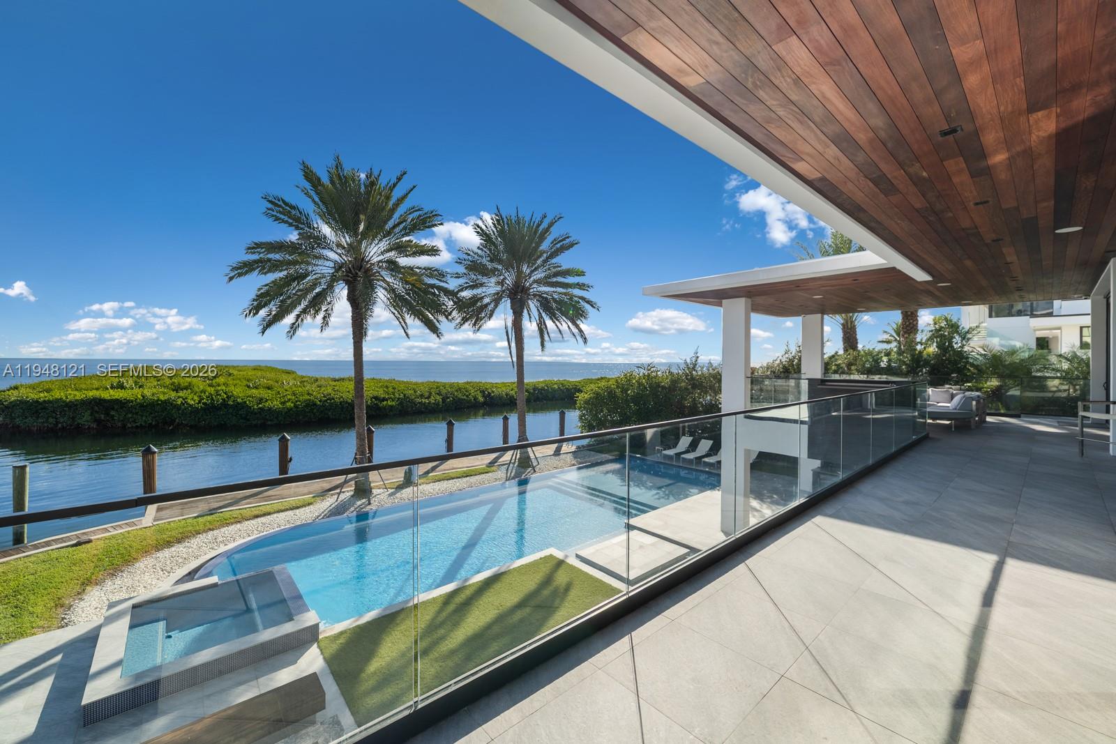 A modern patio features a glass-railed infinity pool overlooking a canal and palm trees — 13009 Mar St, Coral Gables FL 33156, CORAL GABLES, FL
