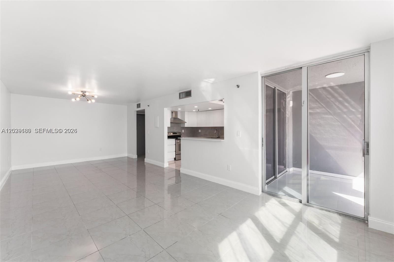 An open-concept living area features light-colored tile flooring, a kitchen pass-through, and a glass balcony door — 17890 W Dixie Hwy 501 Hwy # 501, North Miami Beach FL 33160, NORTH MIAMI BEACH, FL