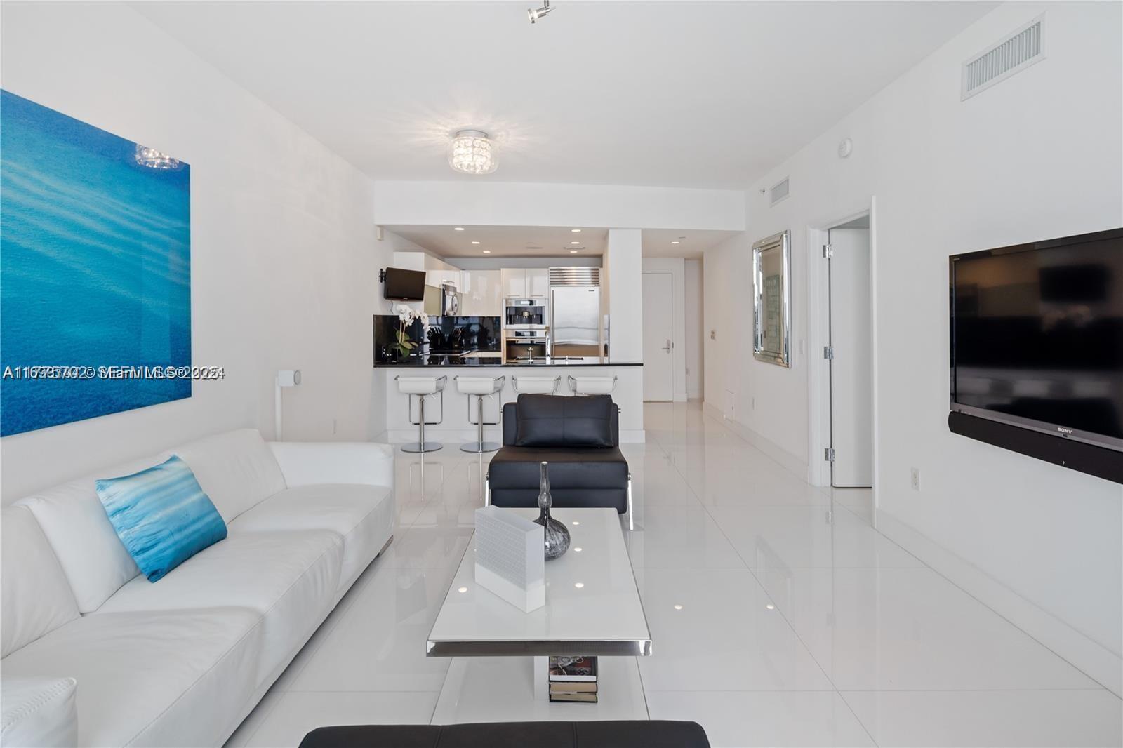 A modern living room features white tile floors, a white sofa, and an open kitchen — 17121 Collins Ave # 1703, Sunny Isles Beach FL 33160, SUNNY ISLES BEACH, FL
