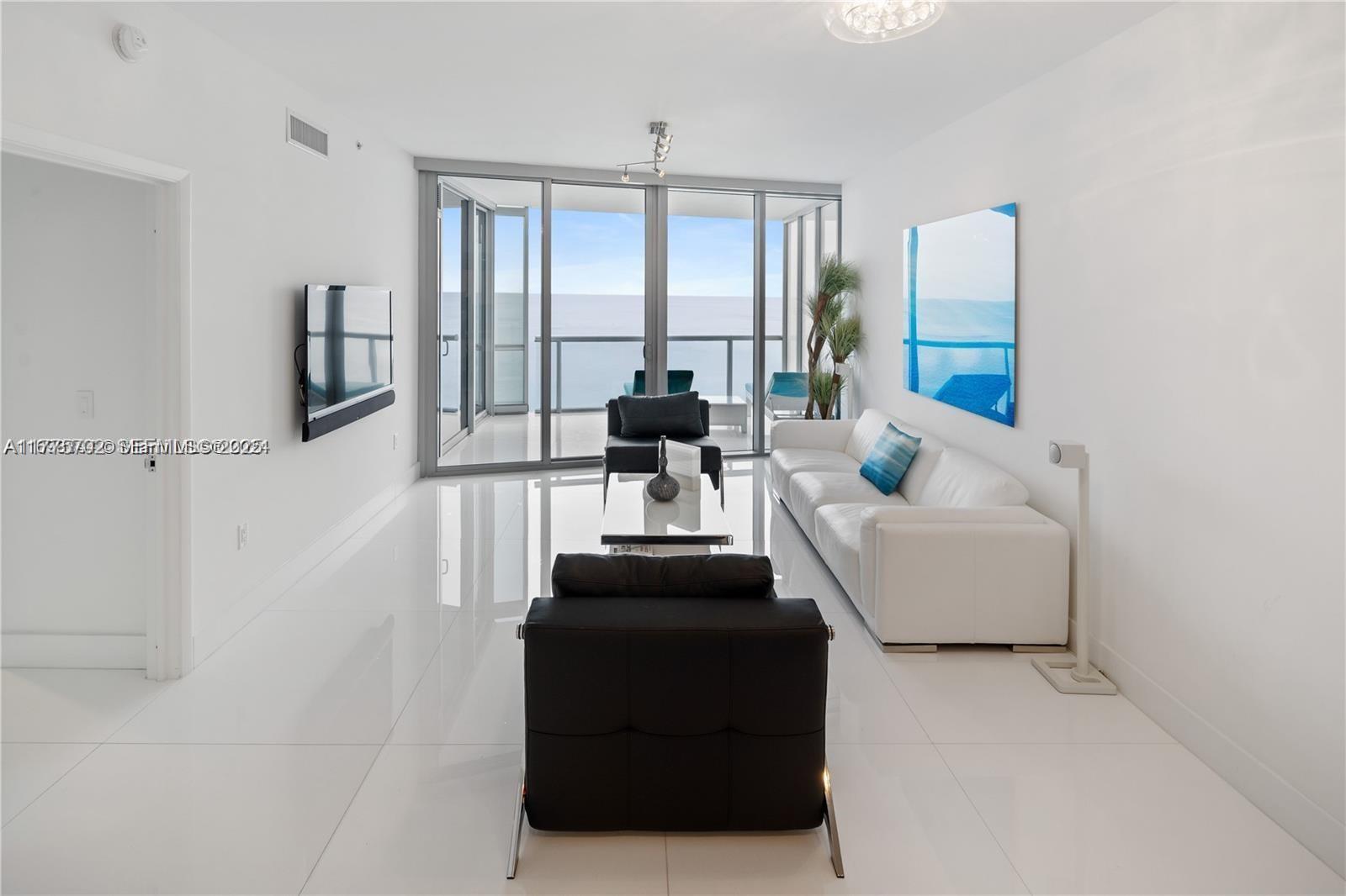 A modern living room features white tile floors, a white sofa, and floor-to-ceiling balcony doors — 17121 Collins Ave # 1703, Sunny Isles Beach FL 33160, SUNNY ISLES BEACH, FL