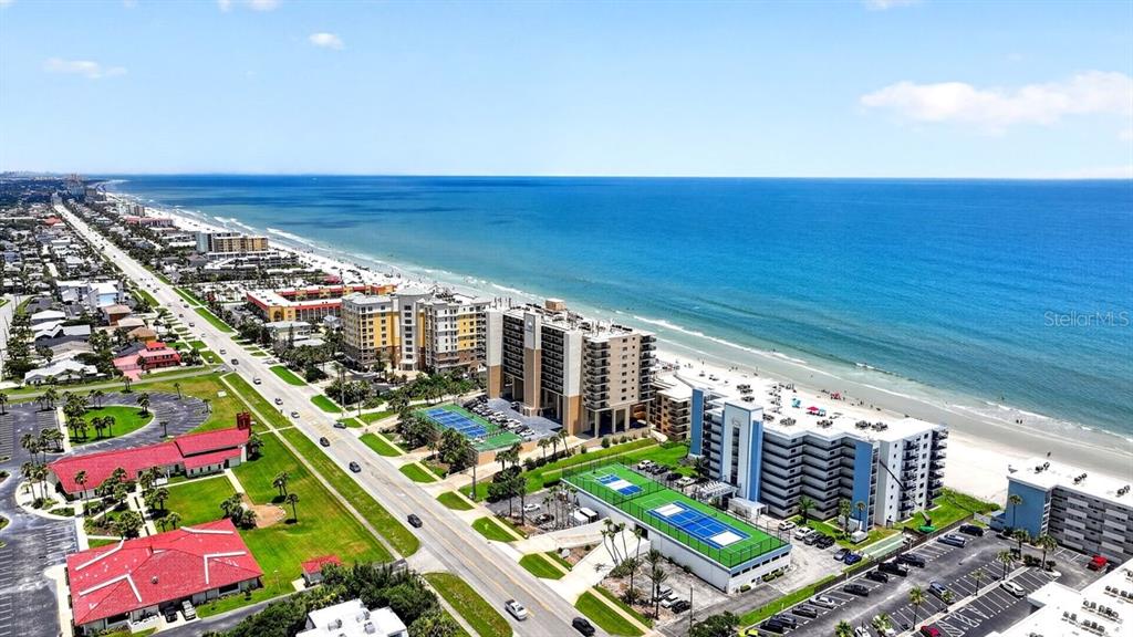 An aerial view shows beachfront condominiums, tennis courts, and a road along the coast — 4139 S ATLANTIC AVENUE UNIT B409, NEW SMYRNA BEACH, FL