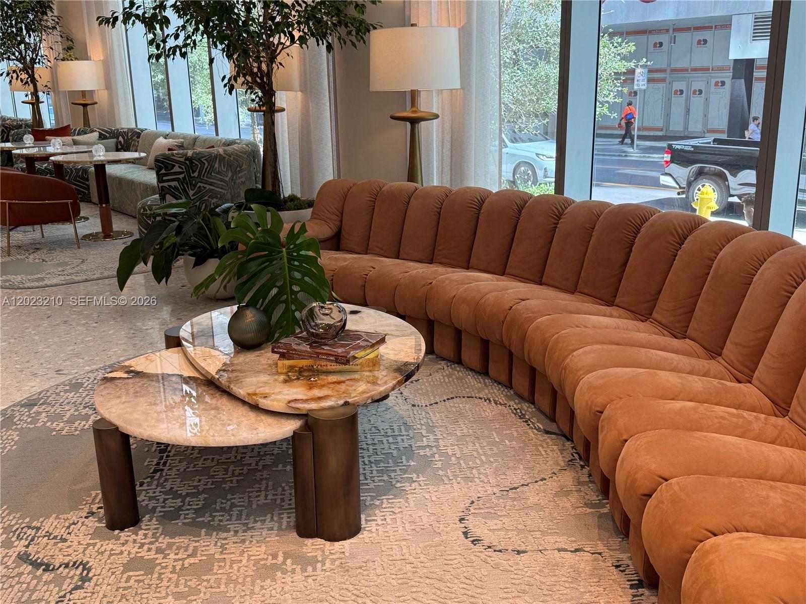 A modern lounge area features a curved tan sofa, stone coffee tables, and large windows — 77 SE 5th ST # 2111, Miami FL 33131, MIAMI, FL