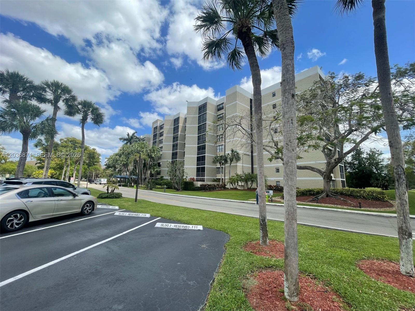 A multi-story residential building stands behind a parking lot with palm trees and landscaping — 16300 Golf Club Rd # 508, Weston FL 33326, WESTON, FL