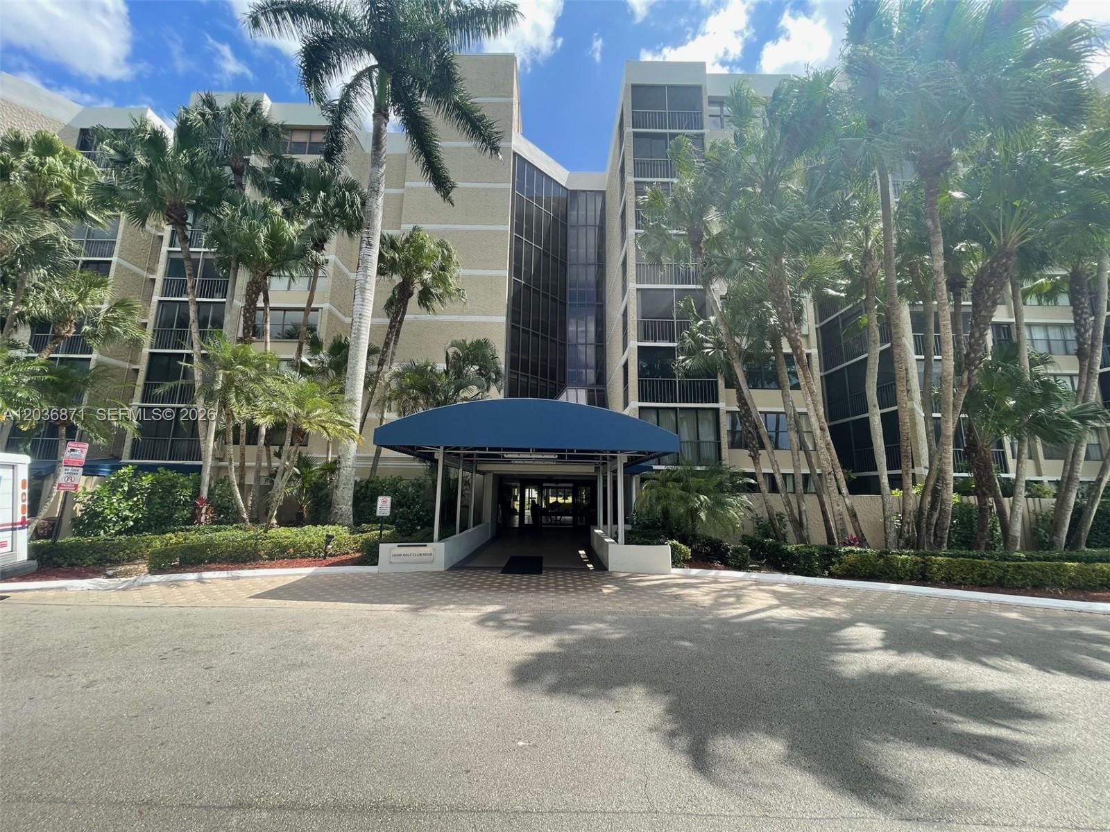 A multi-story residential building features a blue canopy over the entrance and surrounding palm trees — 16300 Golf Club Rd # 508, Weston FL 33326, WESTON, FL