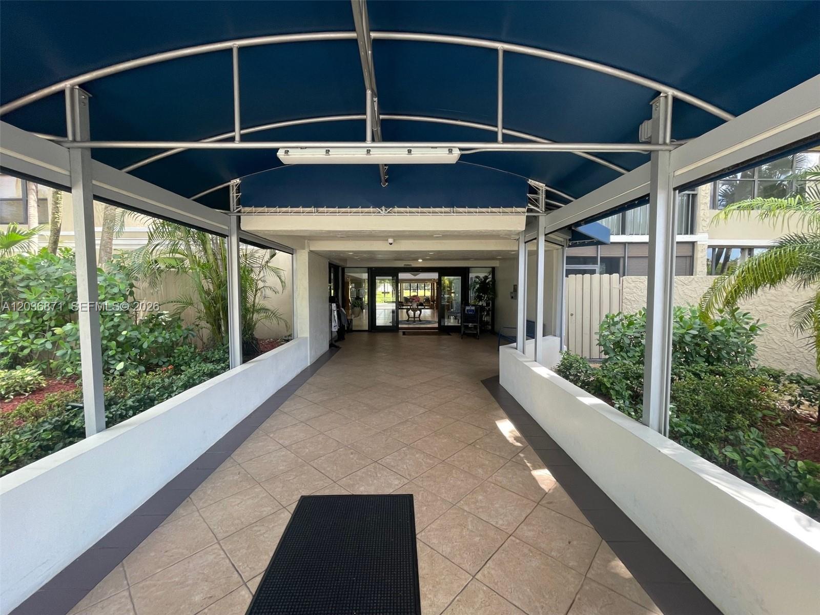 A covered walkway with tiled flooring leads to a building entrance surrounded by lush landscaping — 16300 Golf Club Rd # 508, Weston FL 33326, WESTON, FL