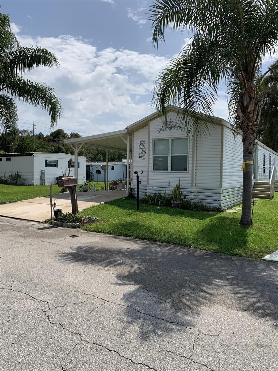 A white manufactured home features a carport, a front lawn, and a palm tree — 3 Mediterranean Boulevard E, Port St. Lucie FL 34952, PORT ST. LUCIE, FL