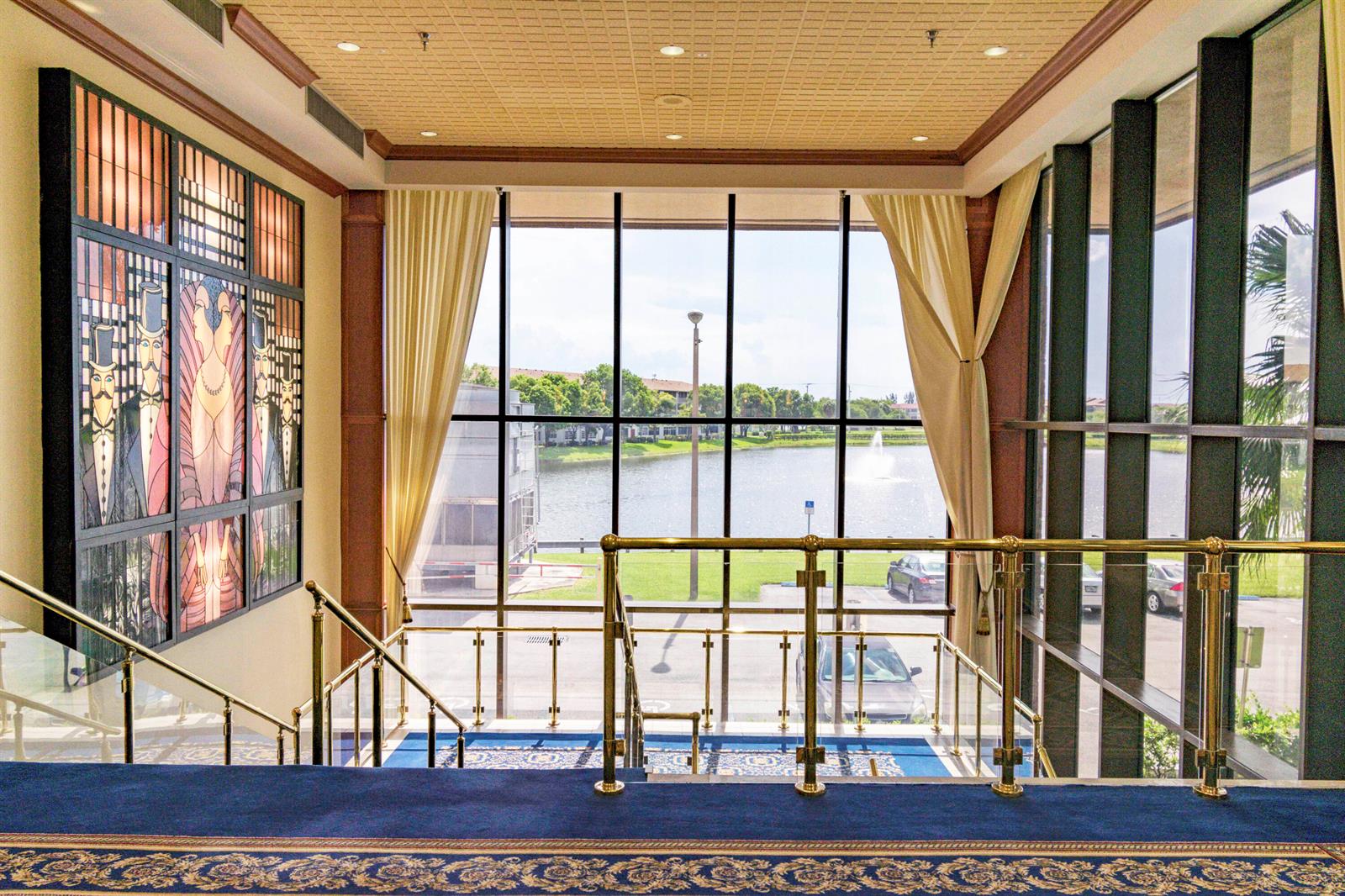 A grand staircase with gold railings overlooks a lake through large floor-to-ceiling windows — 801 SW 141st Avenue # o, Pembroke Pines FL 33027, PEMBROKE PINES, FL