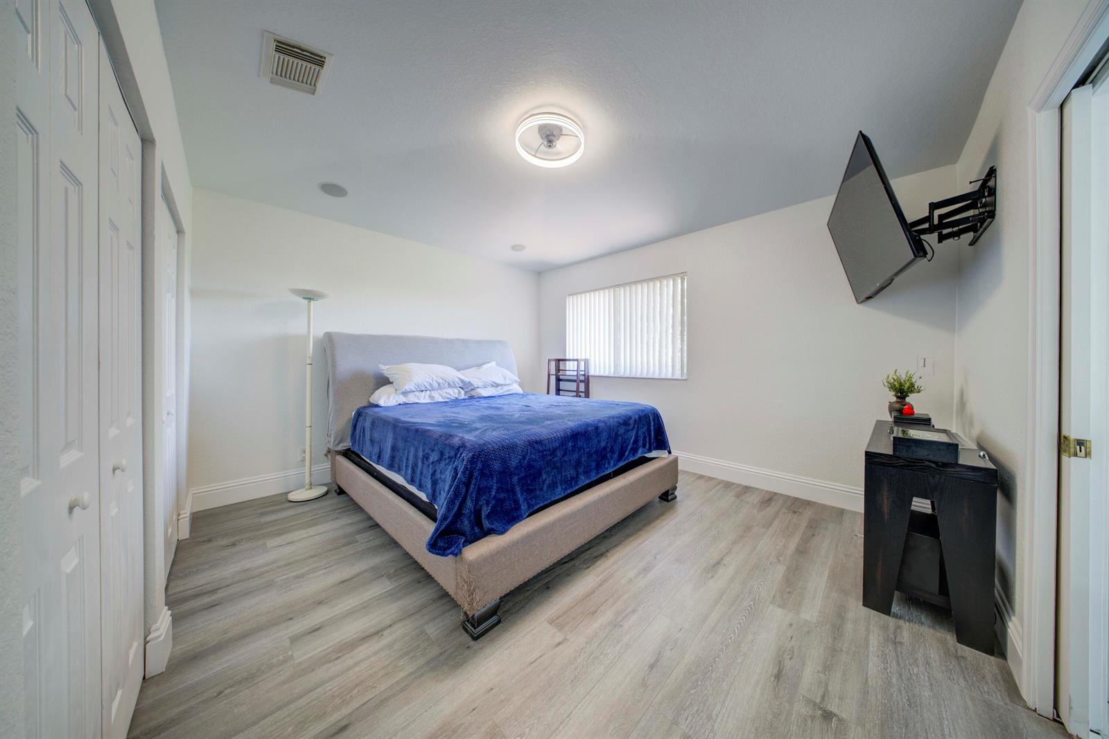 A bedroom features a bed with a blue blanket, wood-look flooring, and a wall-mounted television — 2364 Center Stone Lane, Riviera Beach FL 33404, RIVIERA BEACH, FL