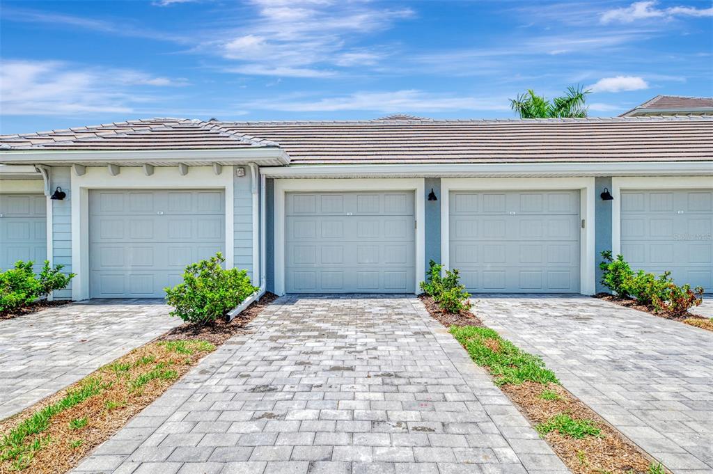 A row of three garage doors with light blue siding and paver driveways under a roof — 12520 GALAPAGOS COURT UNIT 102, VENICE, FL