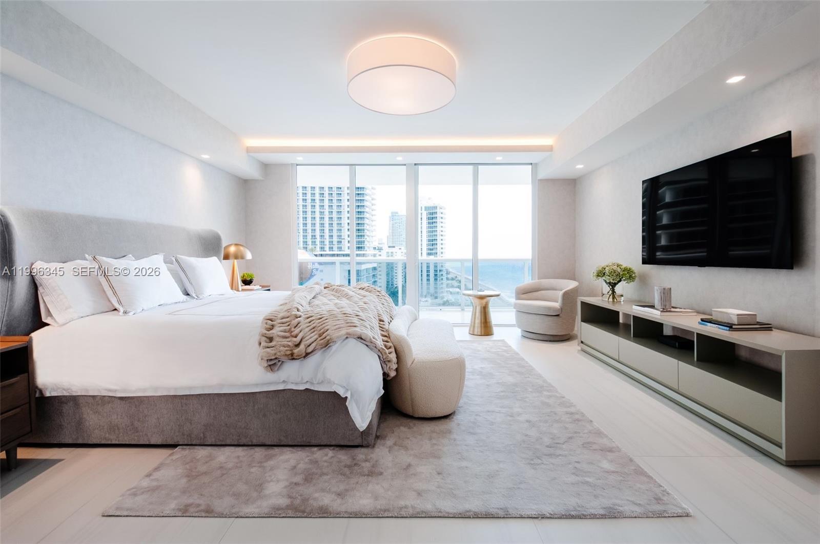 A modern bedroom features a king bed, a wall-mounted television, and floor-to-ceiling windows overlooking buildings — 1830 S Ocean Dr # 1501, Hallandale Beach FL 33009, HALLANDALE BEACH, FL