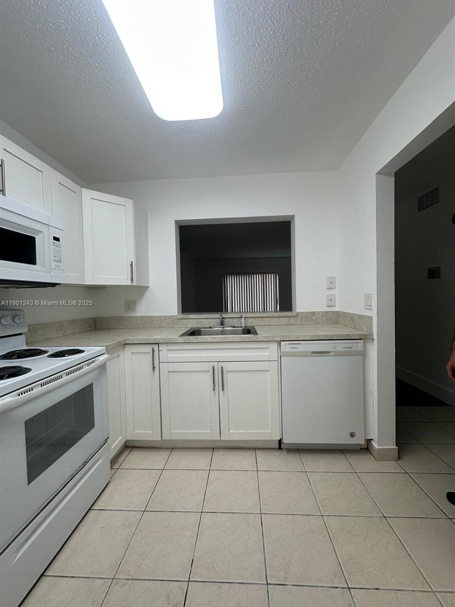 A kitchen features white cabinets, white appliances, a pass-through window, and light-colored tile flooring — 18101 NW 68th Ave # E210, Hialeah FL 33015, HIALEAH, FL