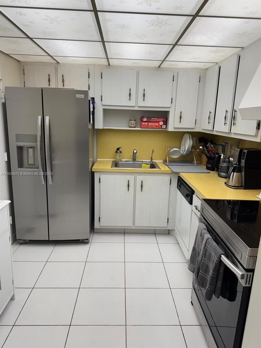 A kitchen features white cabinets, yellow countertops, a stainless steel refrigerator, and white floor tiles — 500 Three Islands Blvd # 908, Hallandale Beach FL 33009, HALLANDALE BEACH, FL