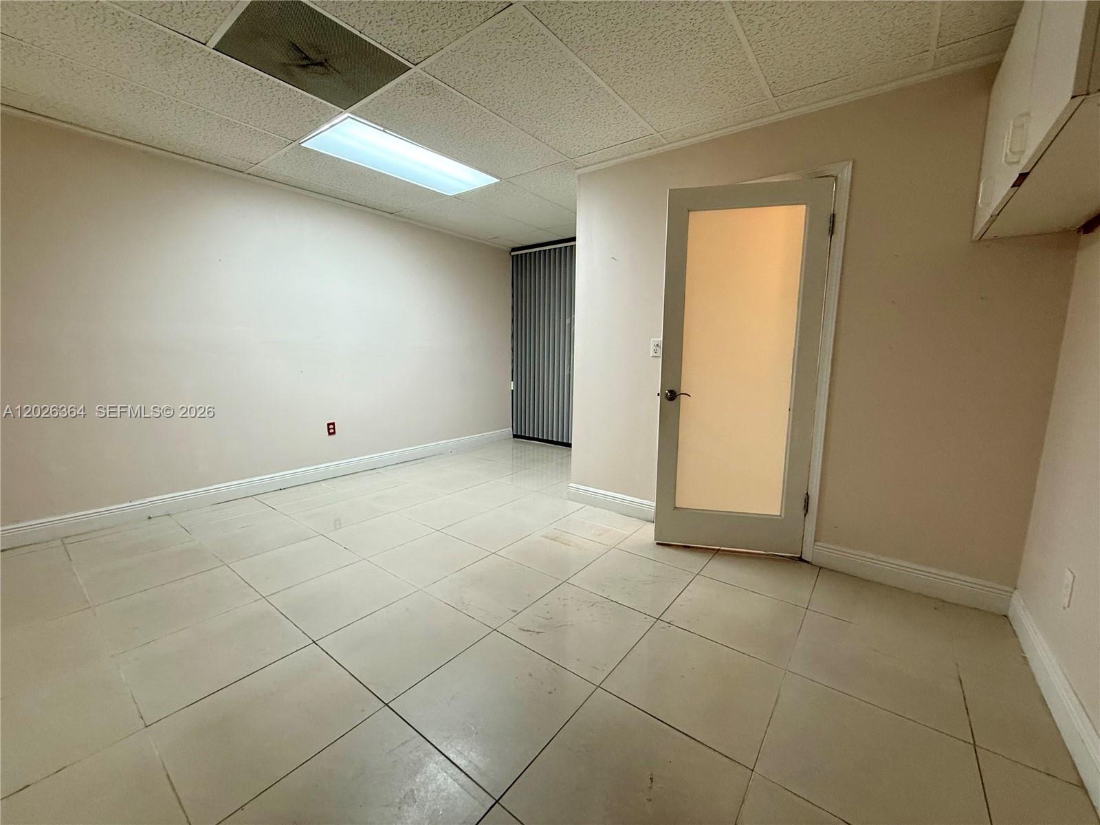 An empty room features light-colored floor tiles, a drop ceiling, and a glass-paneled door — 8370 W Flagler St # 120, Miami FL 33144, MIAMI, FL