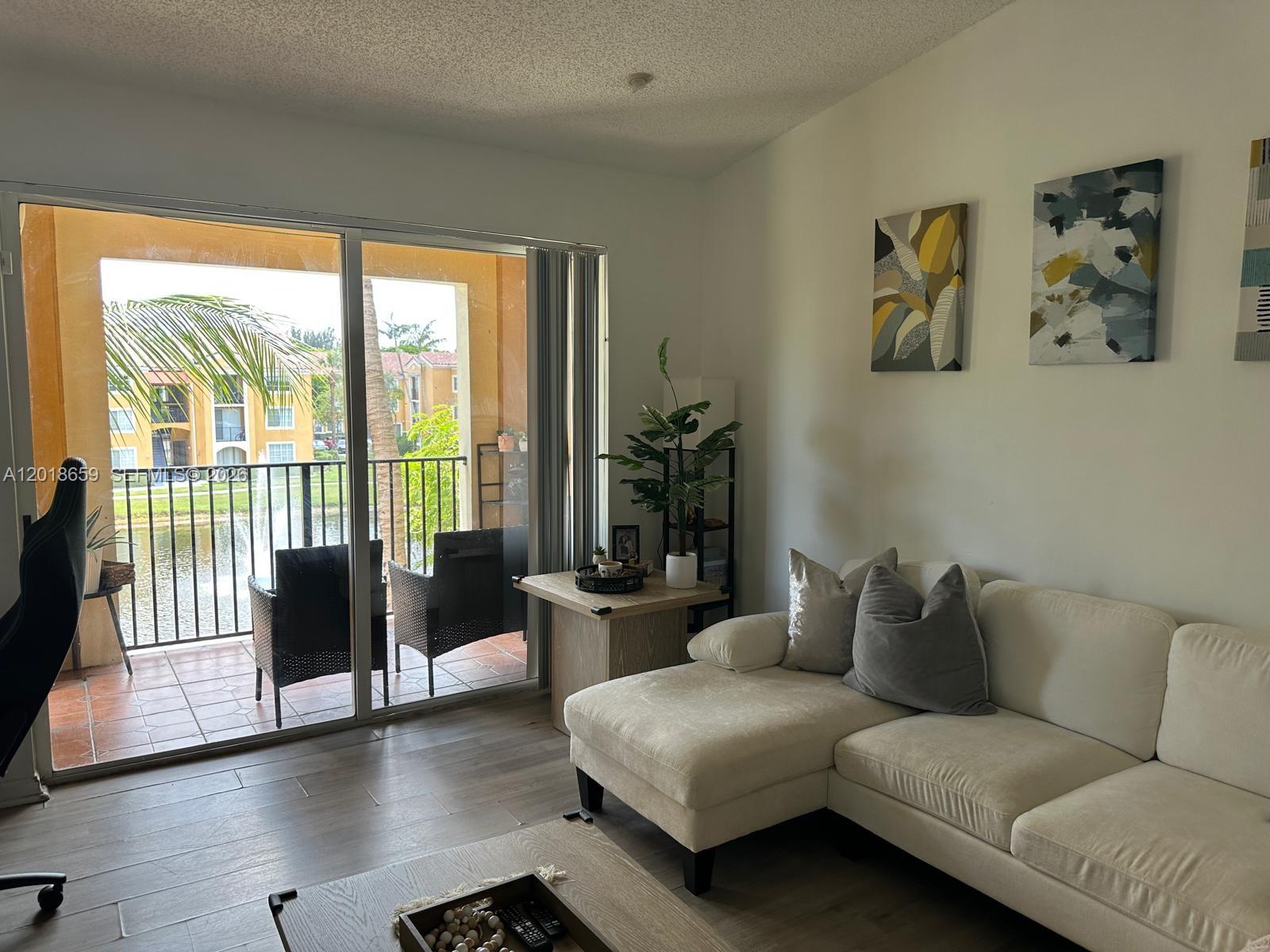 A living room features a white sectional sofa, wood-look flooring, and a balcony overlooking water — 2240 E Preserve Way # 308, Miramar FL 33025, MIRAMAR, FL