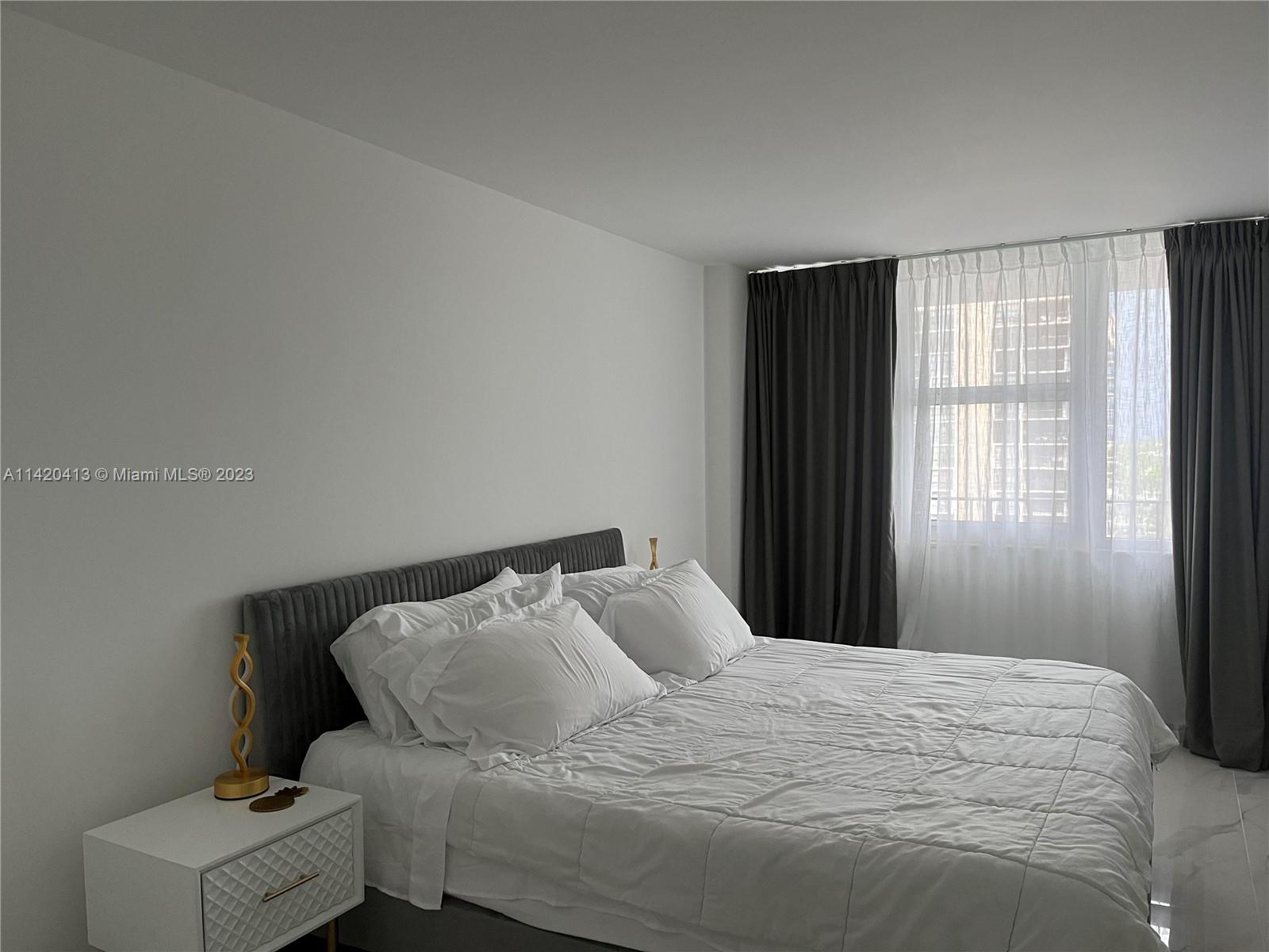 A bedroom features a grey upholstered headboard, white bedding, a white nightstand, and dark curtains — 2030 S Ocean Dr # 1602, Hallandale Beach FL 33009, HALLANDALE BEACH, FL