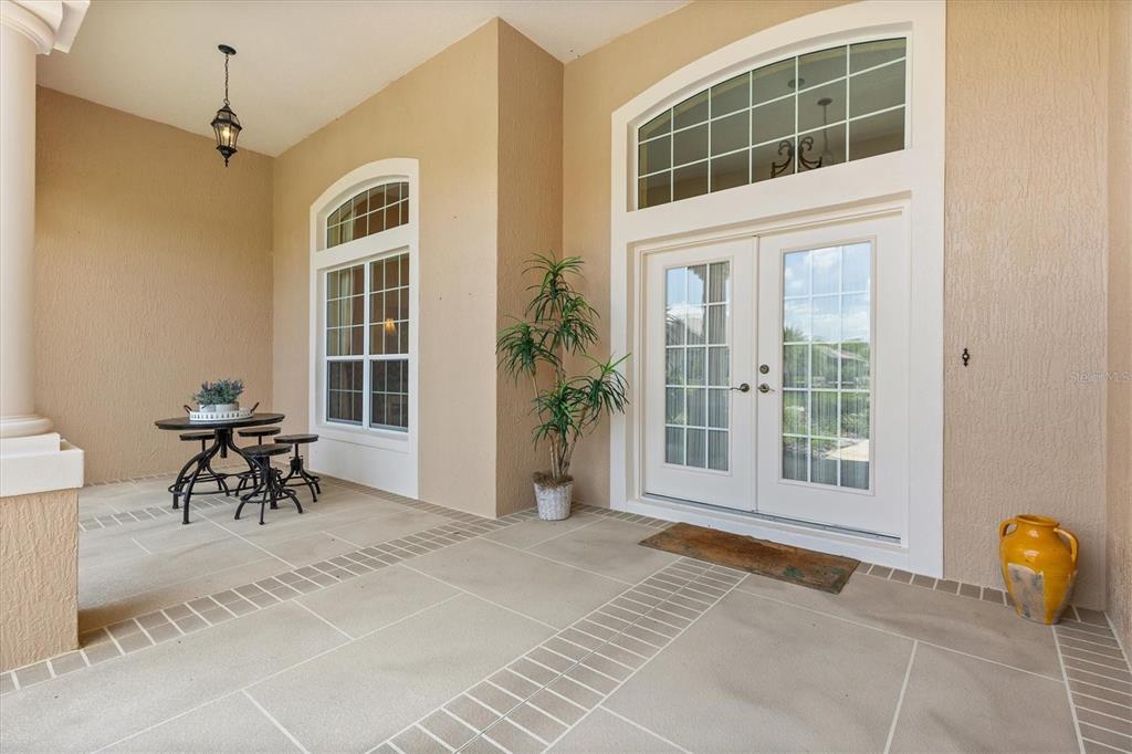 A covered patio features a small bistro table, a potted plant, and double glass doors — 1804 SUNSET RIDGE DRIVE, THE VILLAGES, FL