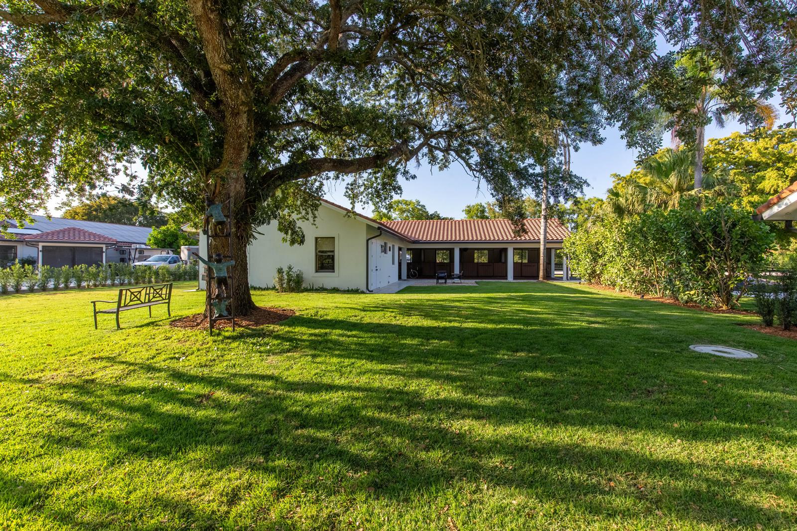 A large oak tree stands in a grassy yard beside a white building with stables — 14828 Rolling Rock Place, Wellington FL 33414, WELLINGTON, FL