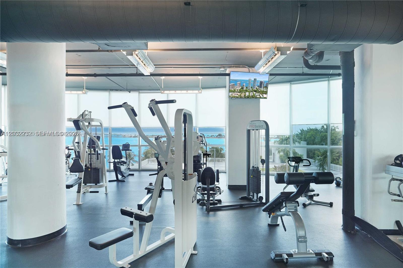 This gym features various exercise machines, white support columns, and floor-to-ceiling windows overlooking the ocean — 2101 Brickell Ave # 2402, Miami FL 33129, MIAMI, FL