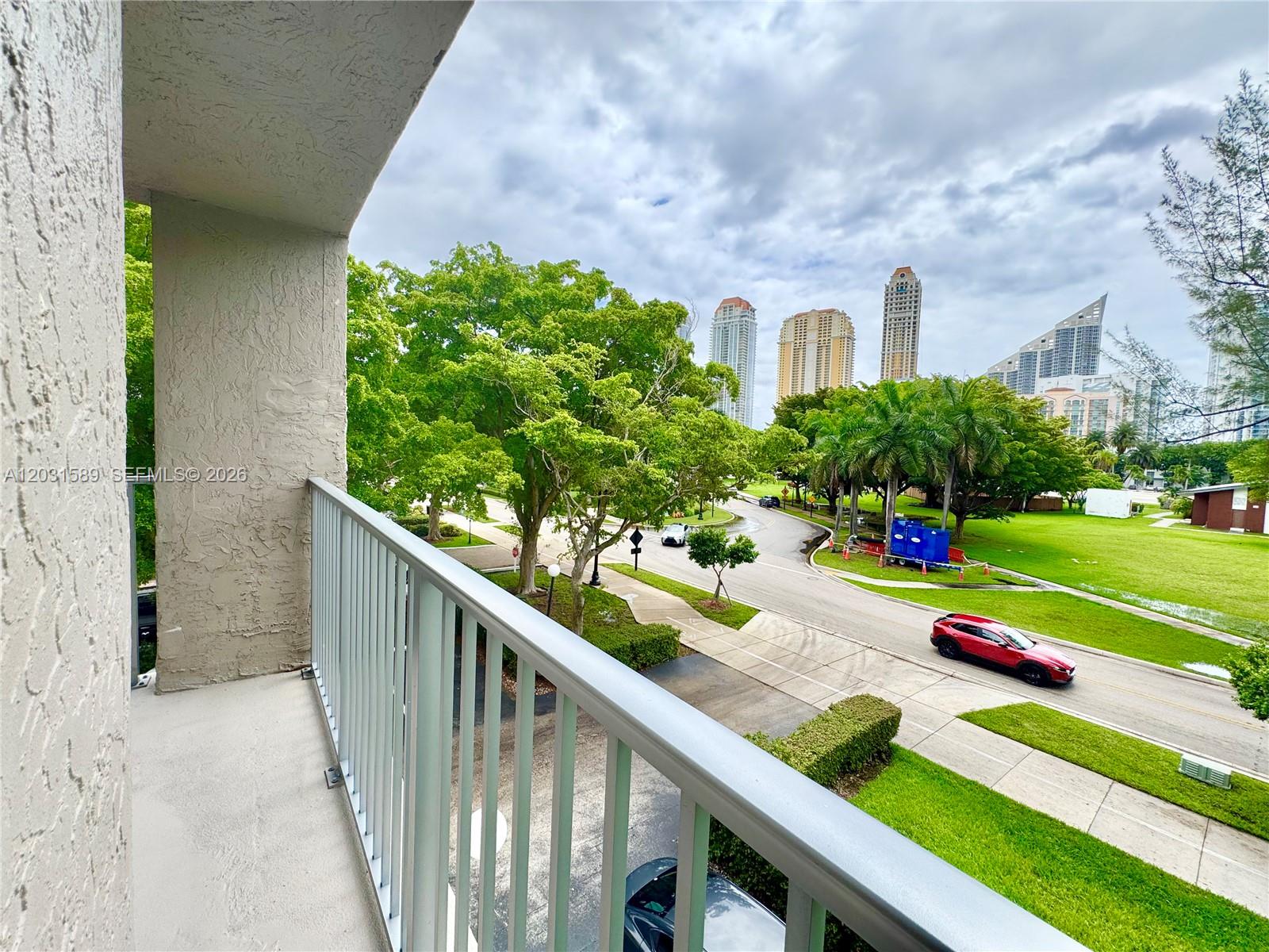 A balcony with a metal railing overlooks a street, green trees, and distant city buildings — 17600 N Bay Rd # N302, Sunny Isles Beach FL 33160, SUNNY ISLES BEACH, FL