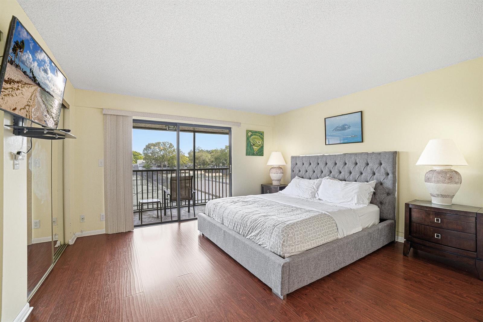 A bedroom features a tufted gray bed, wood-look flooring, and a sliding door to a balcony — 105 Deer Creek Road # 202, Deerfield Beach FL 33442, DEERFIELD BEACH, FL