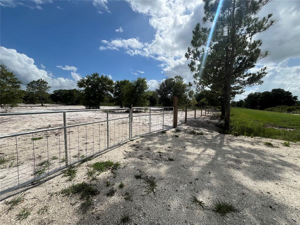 A metal gate opens onto a sandy, fenced lot with scattered trees under a blue sky — 16916 NW 262ND STREET, OKEECHOBEE, FL