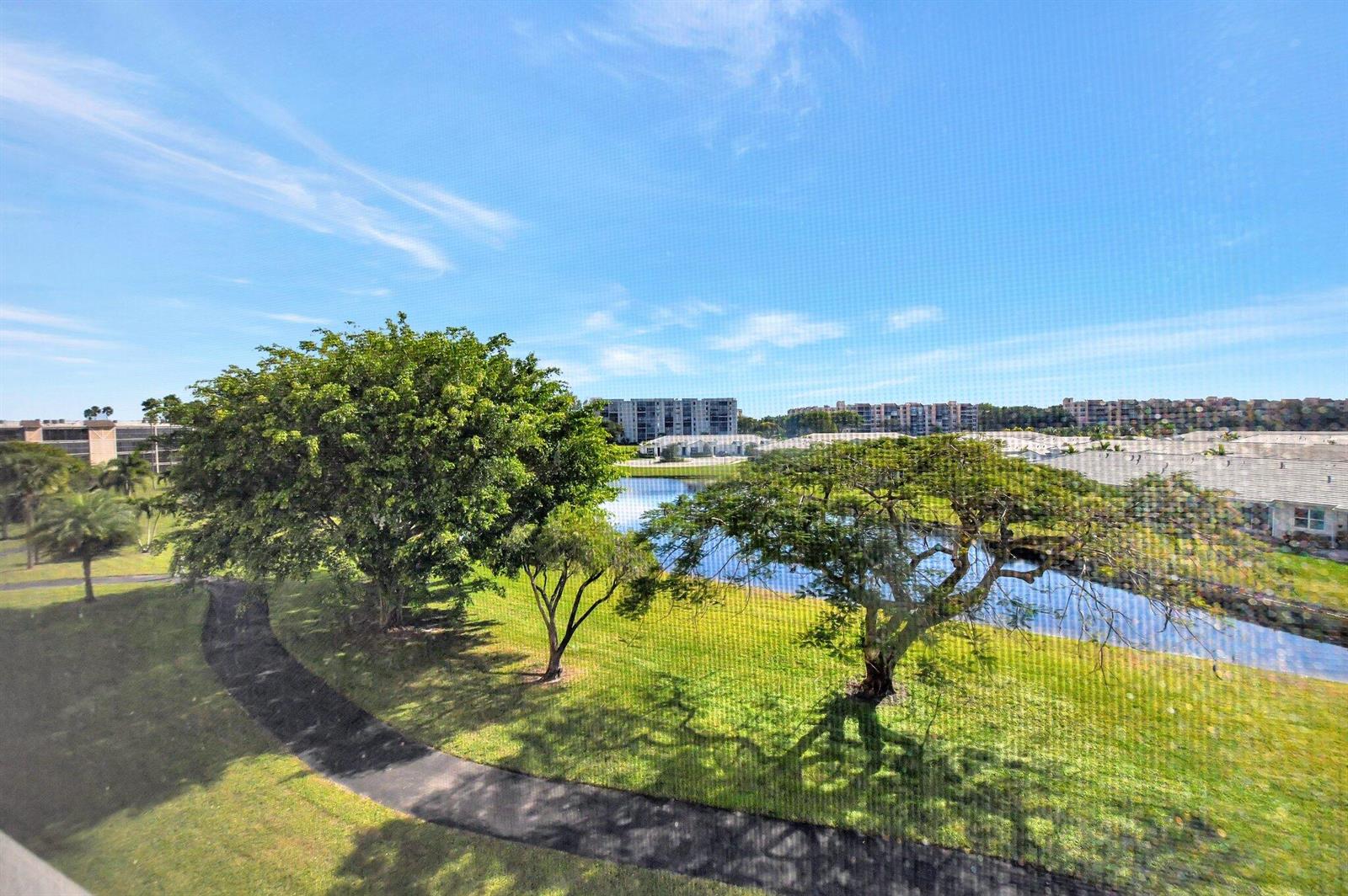 A view of a pond, green lawn, and trees from behind a window screen — 7076 Huntington Lane # 407, Delray Beach FL 33446, DELRAY BEACH, FL