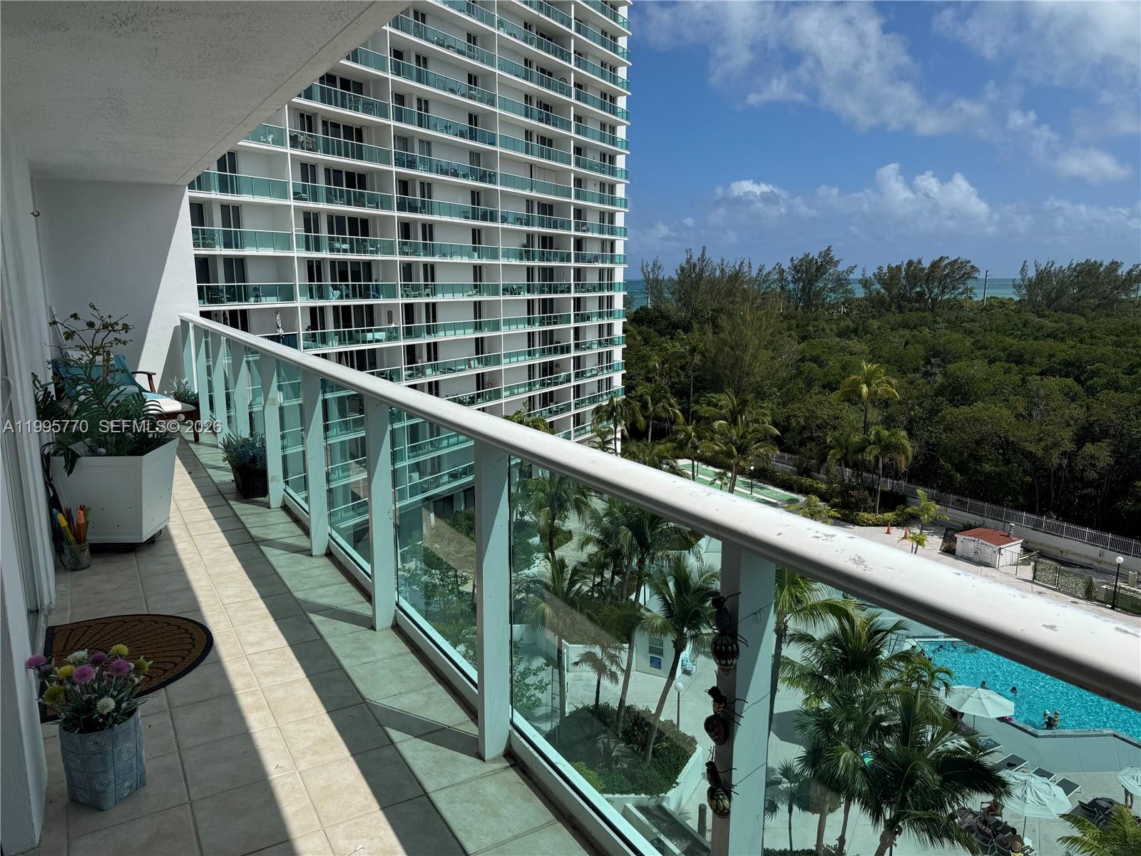A balcony with a glass railing overlooks a swimming pool, palm trees, and a high-rise building — 100 Bayview Dr # 725, Sunny Isles Beach FL 33160, SUNNY ISLES BEACH, FL
