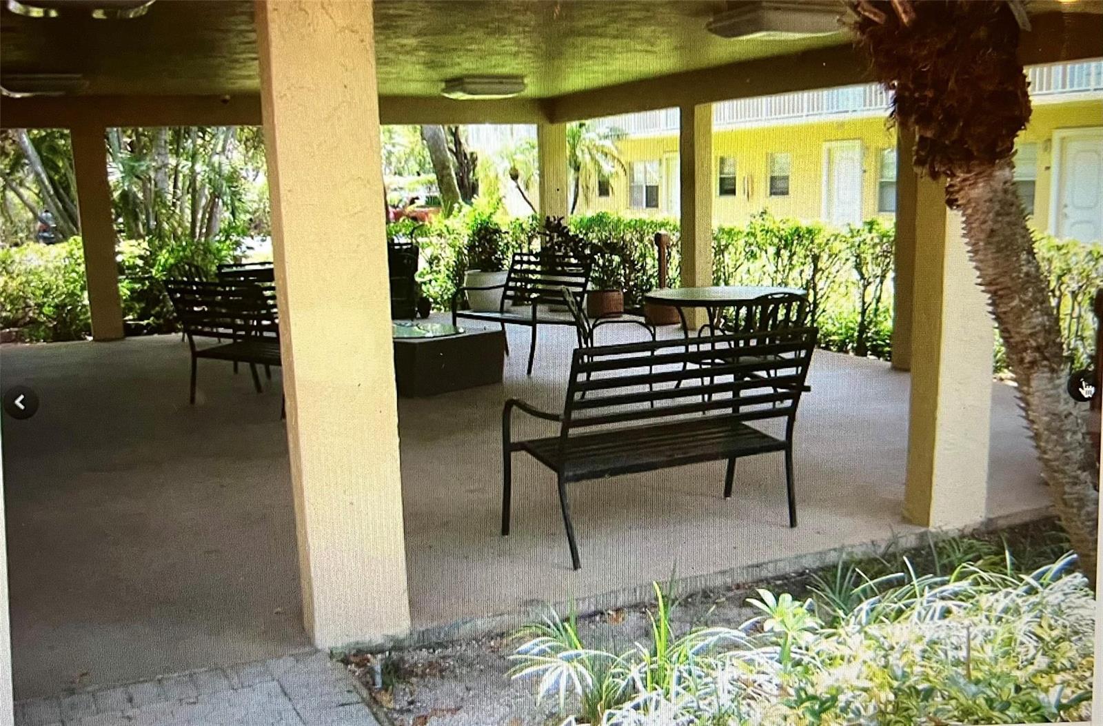A covered outdoor patio features metal benches and tables surrounded by lush green landscaping — 5150 NE 6th Avenue # 130, Oakland Park FL 33334, OAKLAND PARK, FL