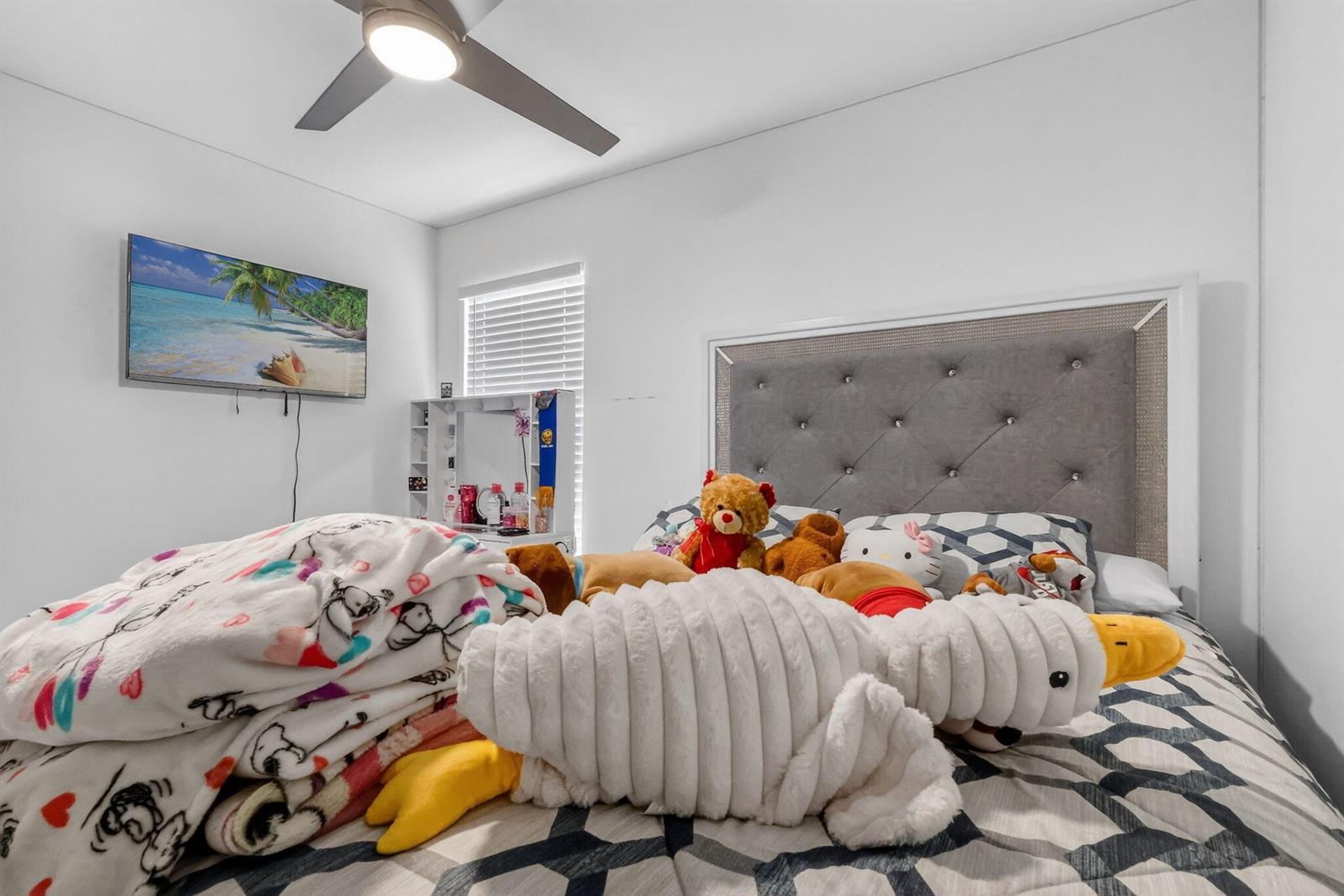 A bedroom features a tufted gray headboard, a ceiling fan, and a bed covered in plush toys — 1691 W 26th Court, Riviera Beach FL 33404, RIVIERA BEACH, FL