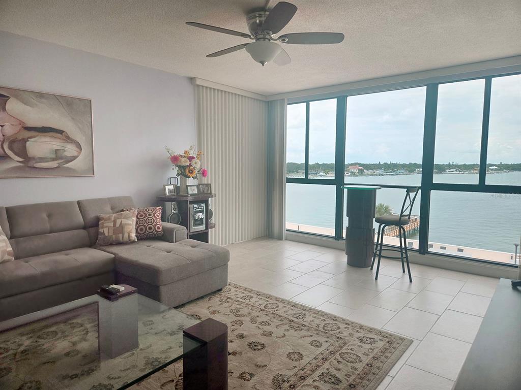 A living room features a grey sectional sofa, tile floors, and floor-to-ceiling windows overlooking water — 7100 SUNSHINE SKYWAY LANE S UNIT 503, ST PETERSBURG, FL