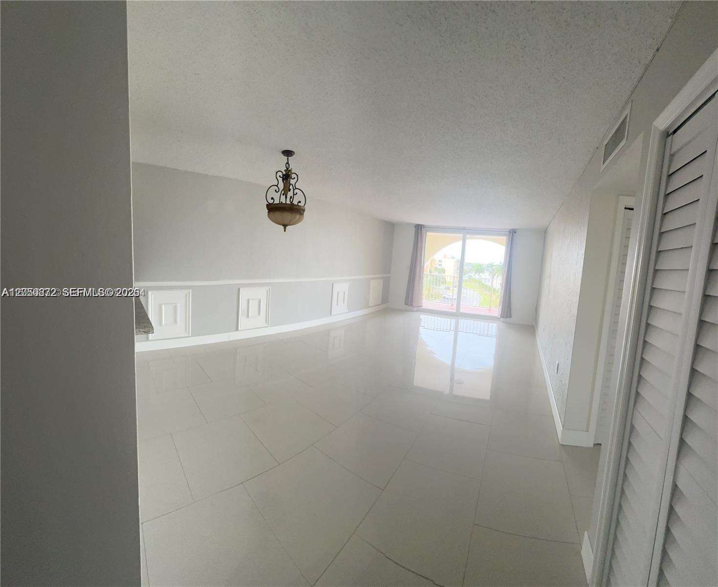 An empty living room features light-colored tile flooring, white wainscoting, and a sliding glass door — 6970 NW 186th St # 502, Hialeah FL 33015, HIALEAH, FL