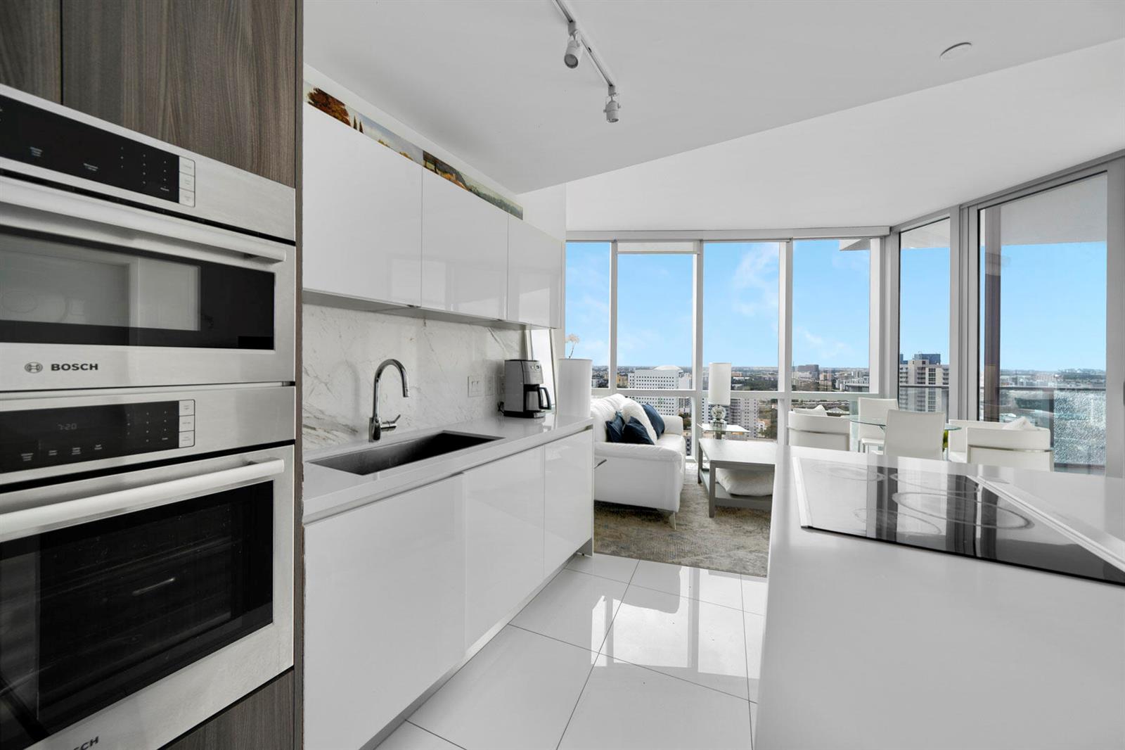 A modern kitchen with white cabinetry and stainless steel appliances overlooks a bright living area — 851 NE 1st Avenue # 2410, Miami FL 33132, MIAMI, FL