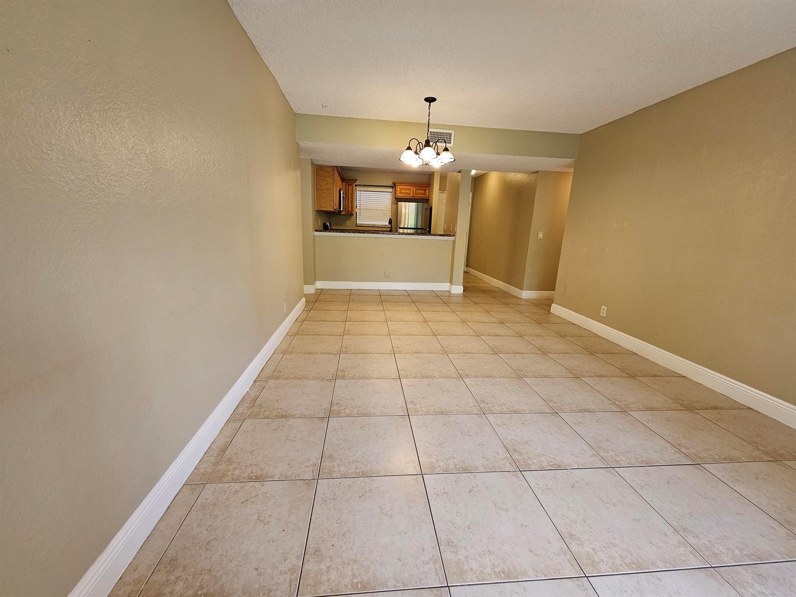 An empty dining area with beige tile flooring leads to an open kitchen with cabinetry — 1461 Windorah Way # C, West Palm Beach FL 33411, WEST PALM BEACH, FL