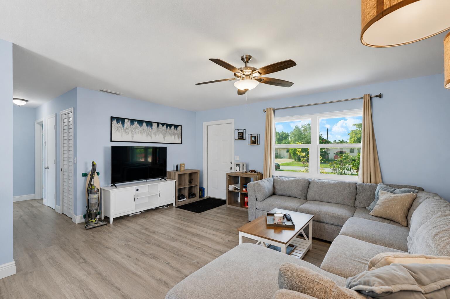 A living room features light blue walls, wood-look flooring, a gray sectional sofa, and a television — 6091 Fair Green Road, West Palm Beach FL 33417, WEST PALM BEACH, FL