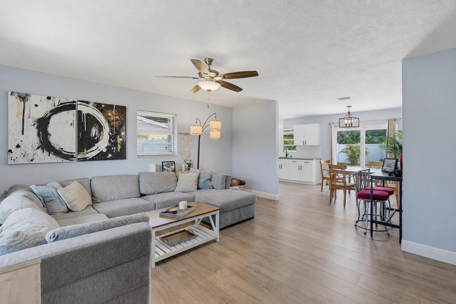 A living room with a gray sectional sofa and wood-look flooring opens to a kitchen — 6091 Fair Green Road, West Palm Beach FL 33417, WEST PALM BEACH, FL