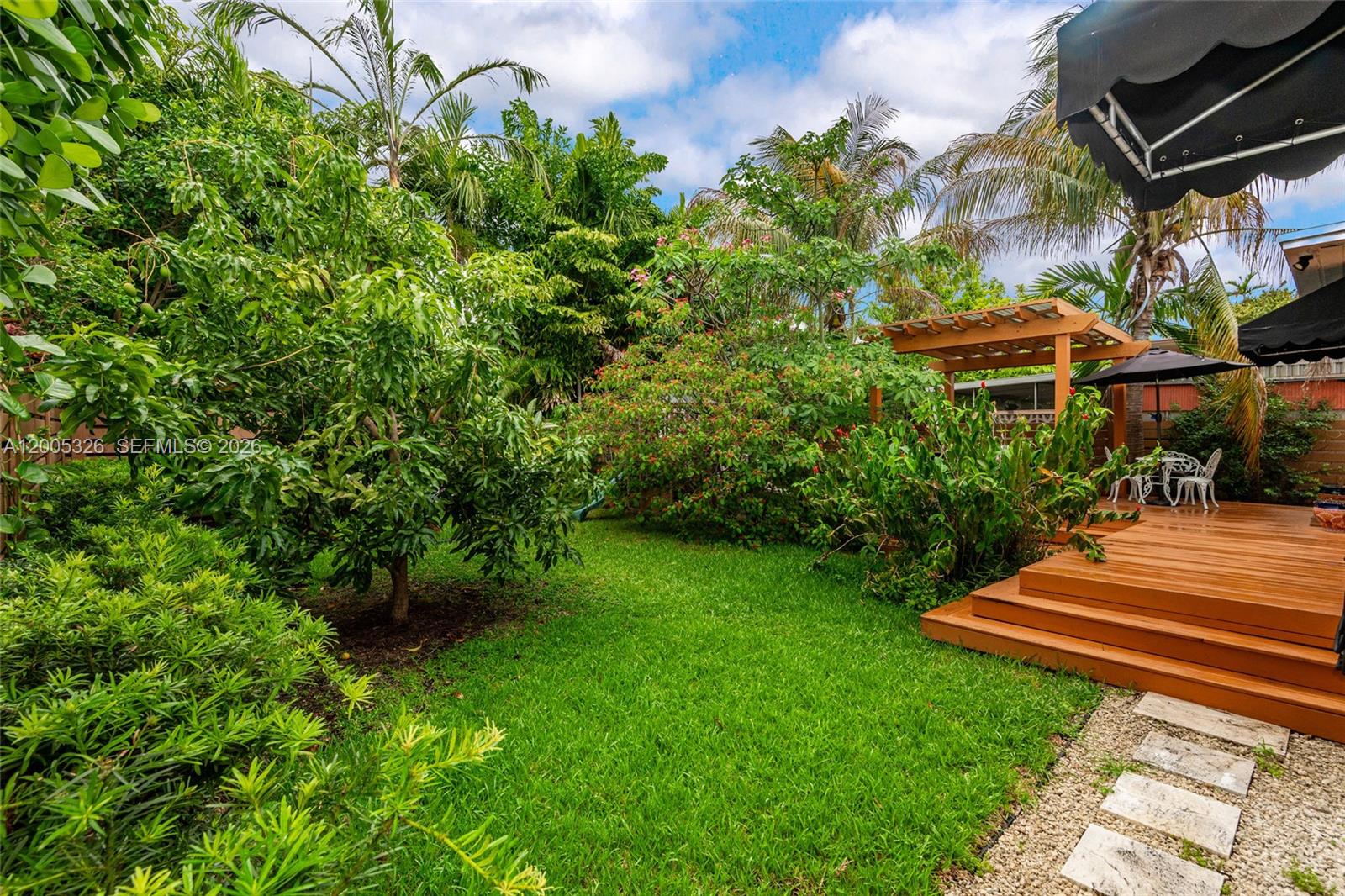 A lush backyard features a wooden deck, a pergola, and various tropical plants and trees — 9048 Carlyle Ave, Surfside FL 33154, SURFSIDE, FL
