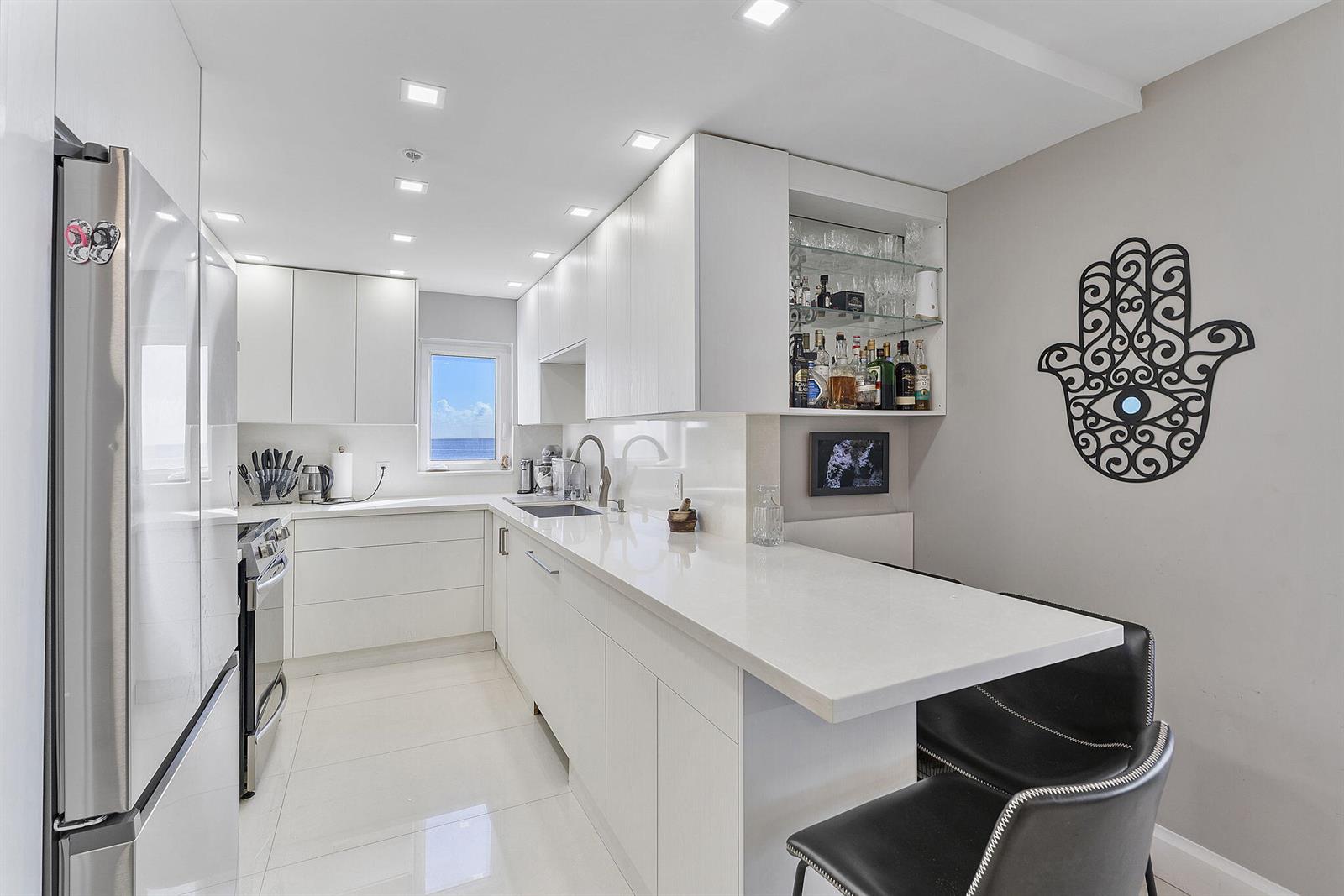 A modern white kitchen features sleek cabinetry, quartz countertops, and a breakfast bar with stools — 4505 S Ocean Boulevard # 307, Highland Beach FL 33487, HIGHLAND BEACH, FL