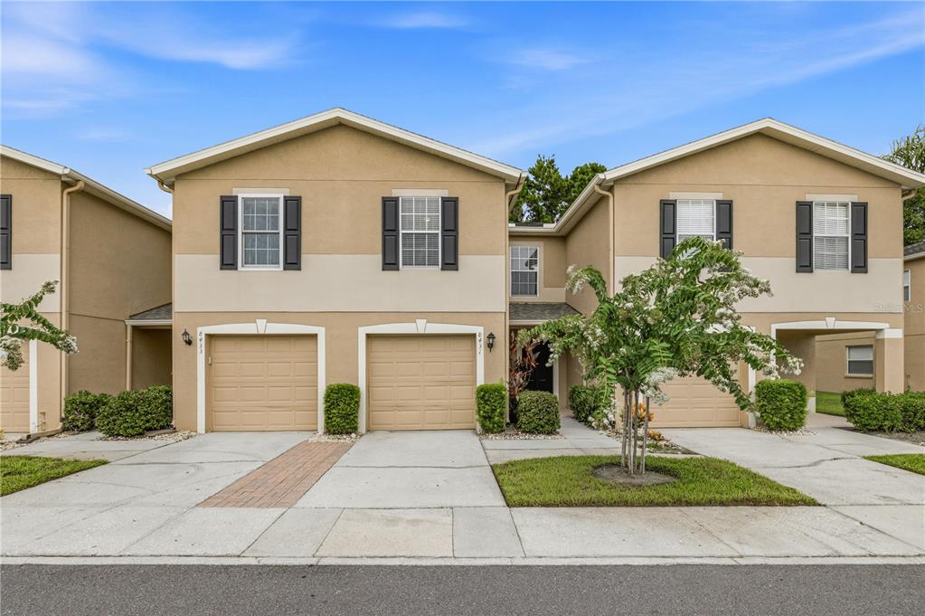 A two-story townhouse exterior with tan stucco walls, two garage doors, and a small tree — 8431 EDGEWATER PLACE BOULEVARD, TAMPA, FL