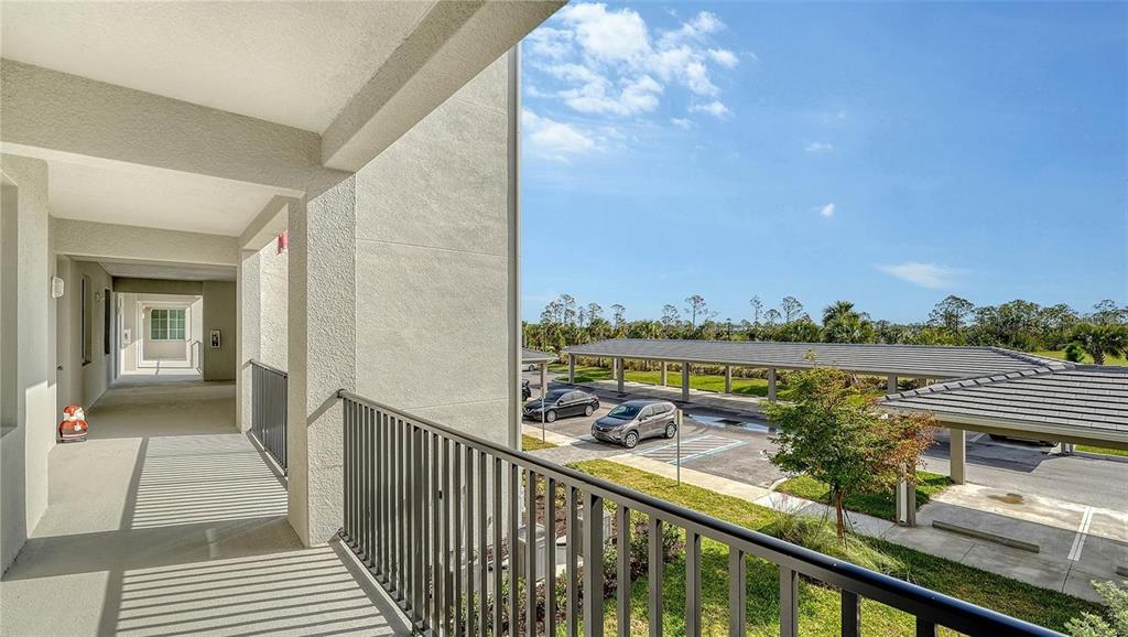 An elevated outdoor balcony overlooks a parking lot with covered carports under a blue sky — 12180 WELLEN GOLF STREET UNIT 203, VENICE, FL