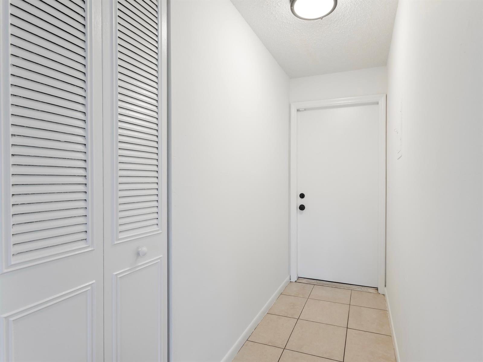 A hallway with white walls, tile flooring, a louvered closet door, and a white door — 450 SW 2nd Avenue # 2050, Boca Raton FL 33432, BOCA RATON, FL