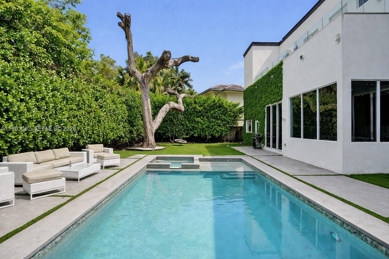 A modern white house features a rectangular swimming pool, patio furniture, and a large tree — 19914 NE 19th Ct, Miami FL 33179, MIAMI, FL