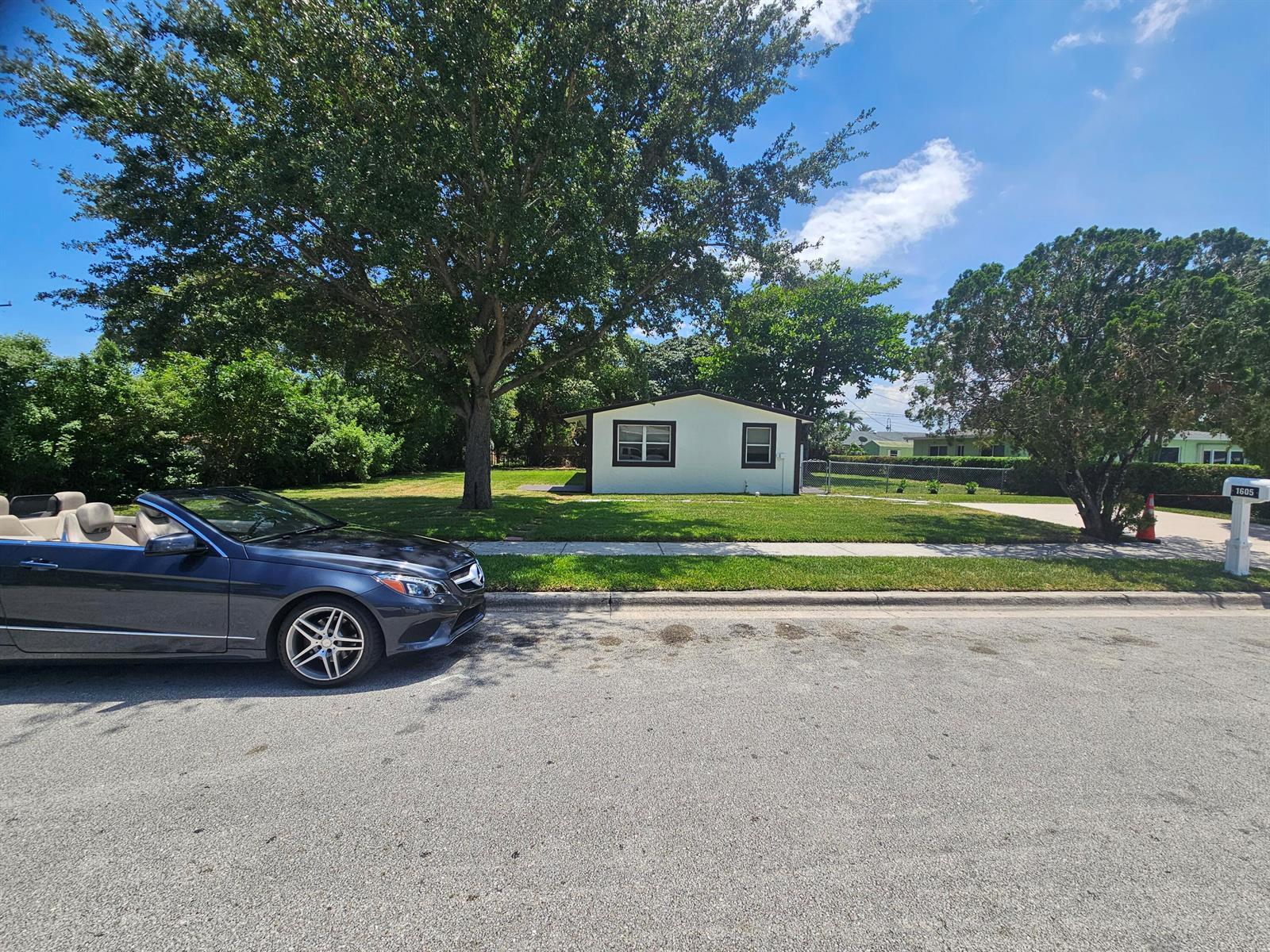 A dark convertible is parked on the street in front of a small, light-colored house — 1605 W 10th Street, Riviera Beach FL 33404, RIVIERA BEACH, FL