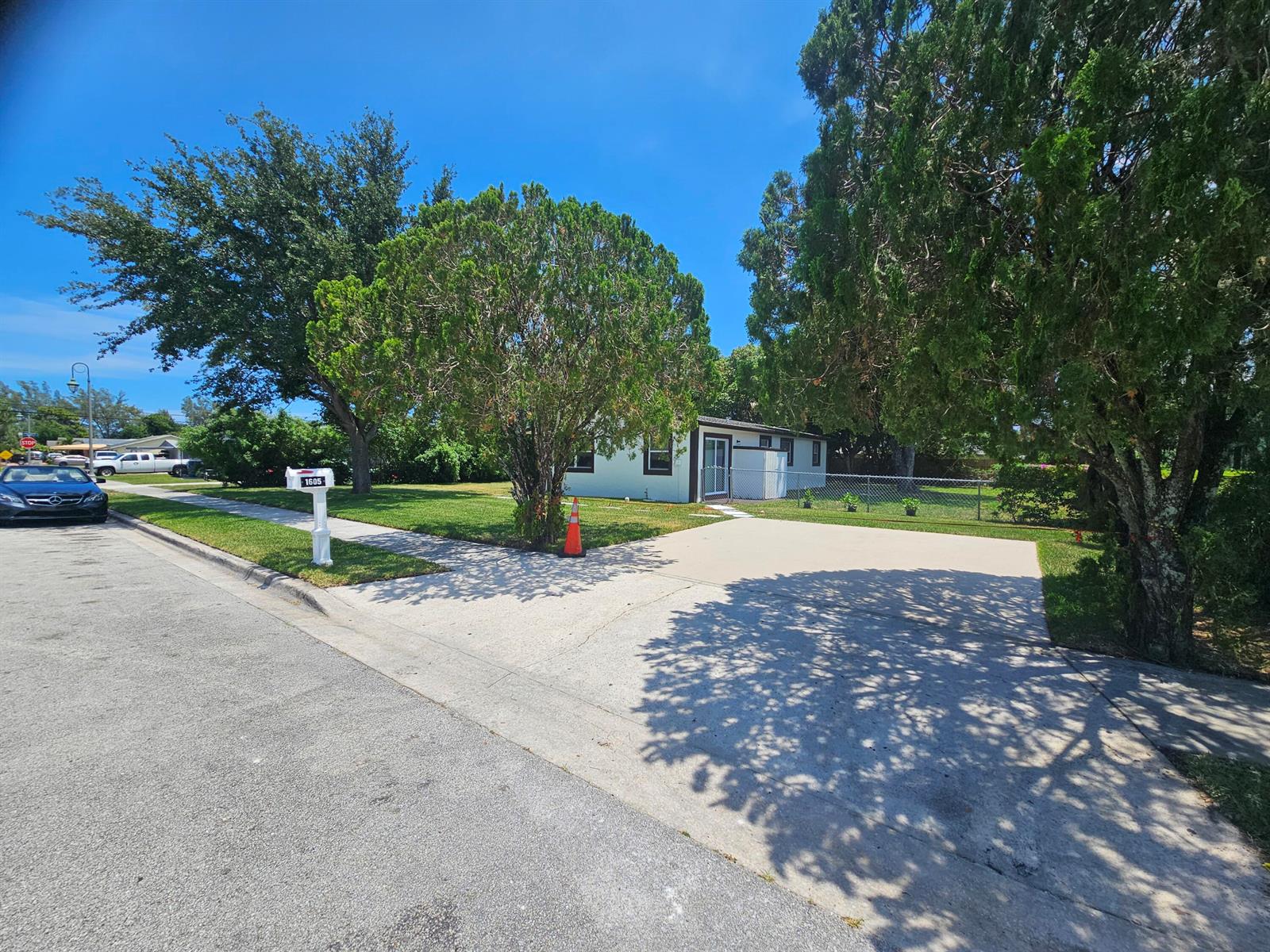 A single-story house with a concrete driveway and a mailbox stands behind large trees — 1605 W 10th Street, Riviera Beach FL 33404, RIVIERA BEACH, FL