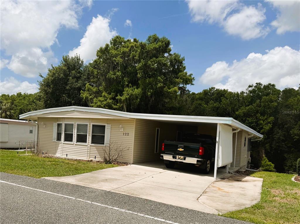 A single-story mobile home with beige siding and a carport featuring a parked pickup truck — 122 PALO VERDE DRIVE, LEESBURG, FL