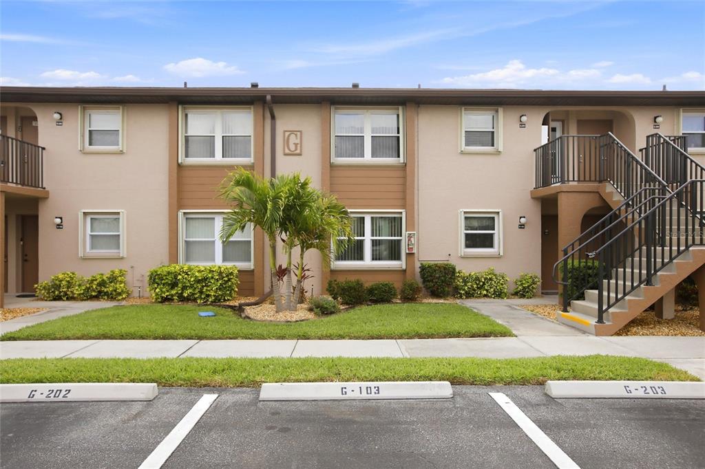 A two-story apartment building exterior features tan stucco walls, brown trim, and assigned parking spaces — 25100 SANDHILL BOULEVARD UNIT G103, PUNTA GORDA, FL