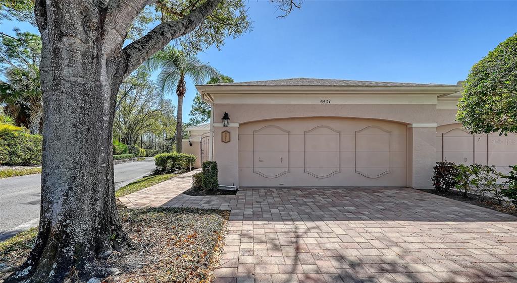 A beige house exterior features a three-car garage and a paved driveway under a tree — 5521 CHANTECLAIRE UNIT 21, SARASOTA, FL