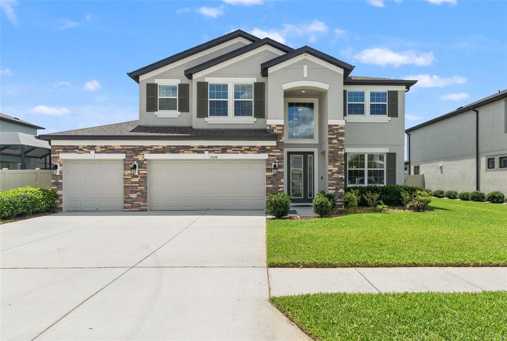 A two-story suburban house features a stone-accented facade, a three-car garage, and a lawn — 7648 SWEETER TIDE TRAIL, WESLEY CHAPEL, FL