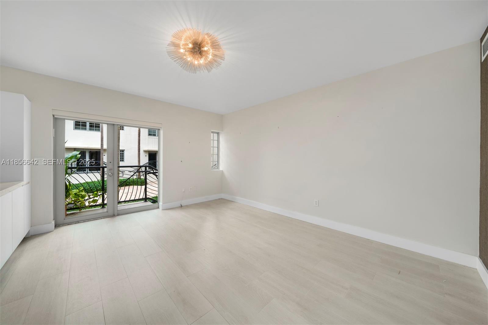 An empty room features light wood-look flooring, a modern chandelier, and a glass balcony door — 435 21st St # 103, Miami Beach FL 33139, MIAMI BEACH, FL