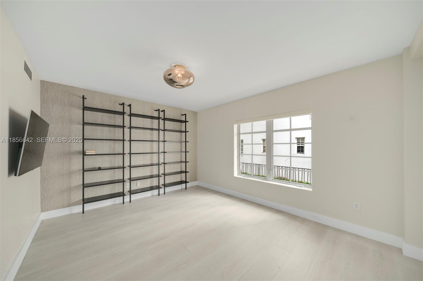 An empty room features light wood flooring, a textured accent wall with shelving, and a window — 435 21st St # 103, Miami Beach FL 33139, MIAMI BEACH, FL