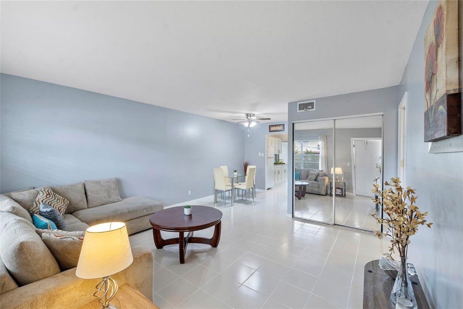 A living room with light blue walls, tile flooring, a sectional sofa, and mirrored closet doors — 7500 NW 5th Court # 104, Margate FL 33063, MARGATE, FL